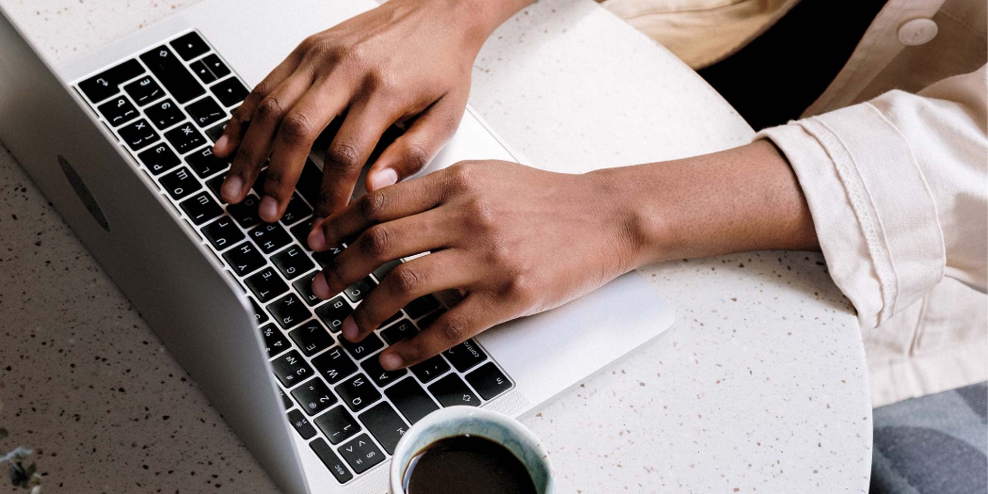 This is an image of a person seated at a table typing on a laptop computer. They are dressed casually and only their hands are visible.