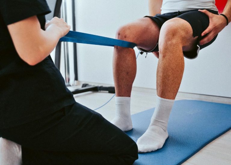 This is an image of a physical therapist working with a patient in a clinic. The patient is in a wall-sit position and the therapist has an elastic band behind the patient's right knee.