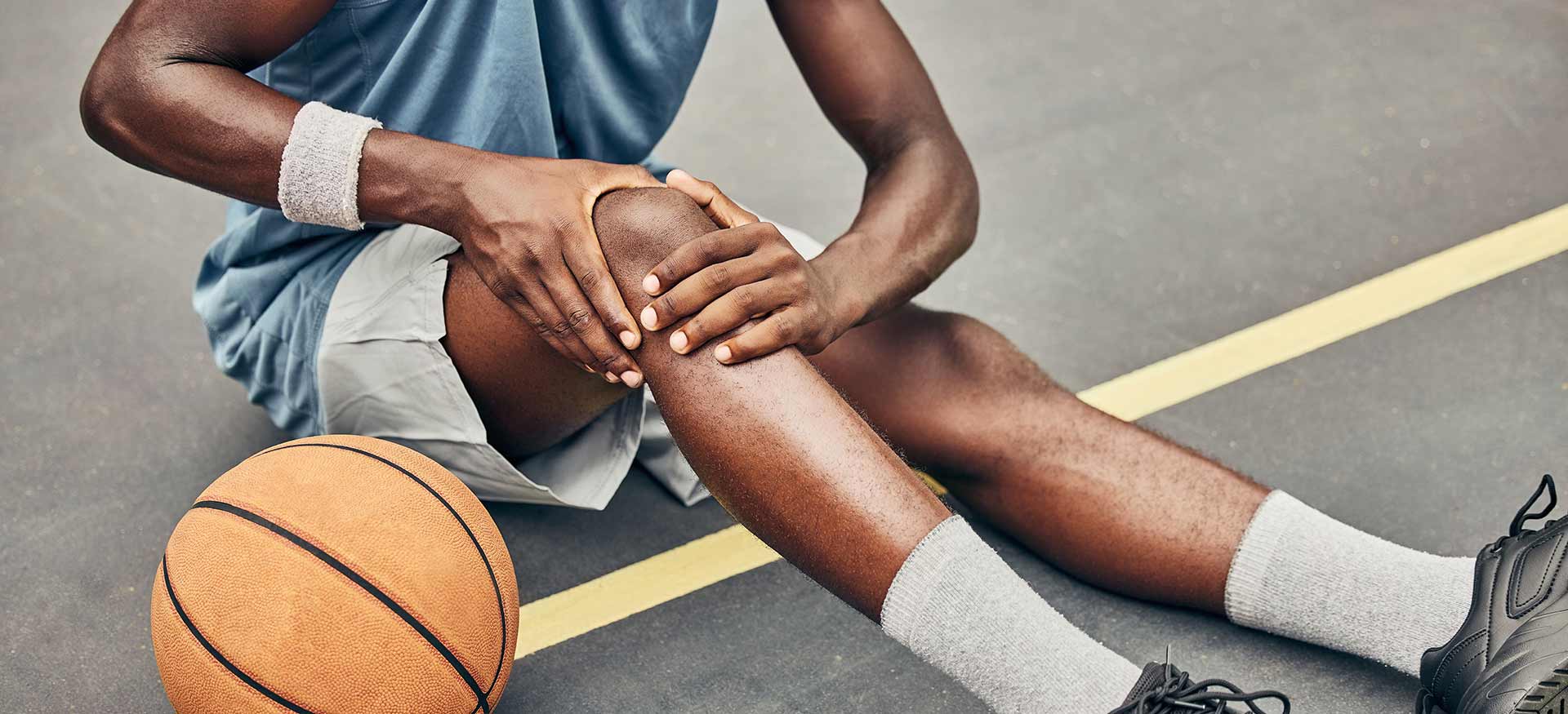 This is an image of a young athletic man sitting on a basketball court holding his right knee, indicating right knee pain or injury.